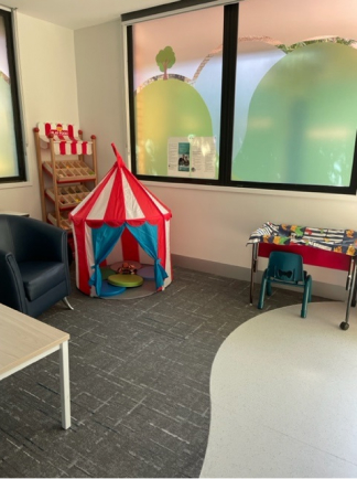 A room with an armchair and table next to a small children's indoor tent. There is an internal window on one wall with colourful tinted glass.