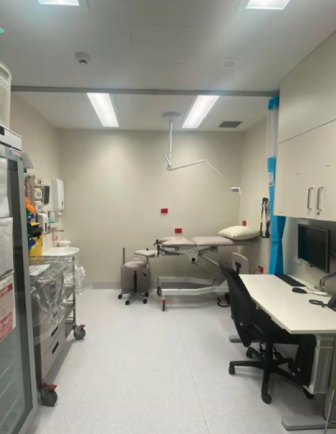 A medical exam room. On the left side there is a fridge and medical equipment. At the back of the room is an examination bed. On the right side there is a desk, computer and chair.